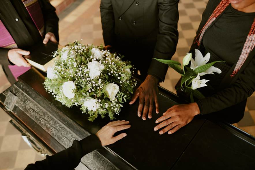 Four individuals touch a dark casket, adorned with white flowers and lilies. The somber scene illustrates loss of sepulcher claims, suggesting grief and legal implications.